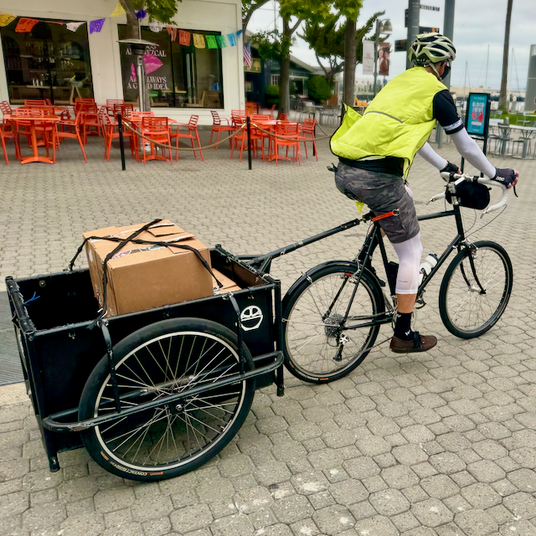 Bicycle Coffee delivery rider transporting boxes of coffee using a bike trailer through a public plaza, wearing a high-visibility vest.