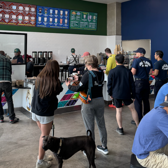 Guests enjoying the space at Bicycle Coffee’s flagship cafe and roastery in Oakland, California, with baristas preparing drinks and colorful menu boards in view.