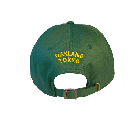 Back of green dad hat with yellow embroidered text reading ‘Oakland Tokyo’ above an adjustable strap with brass buckle.